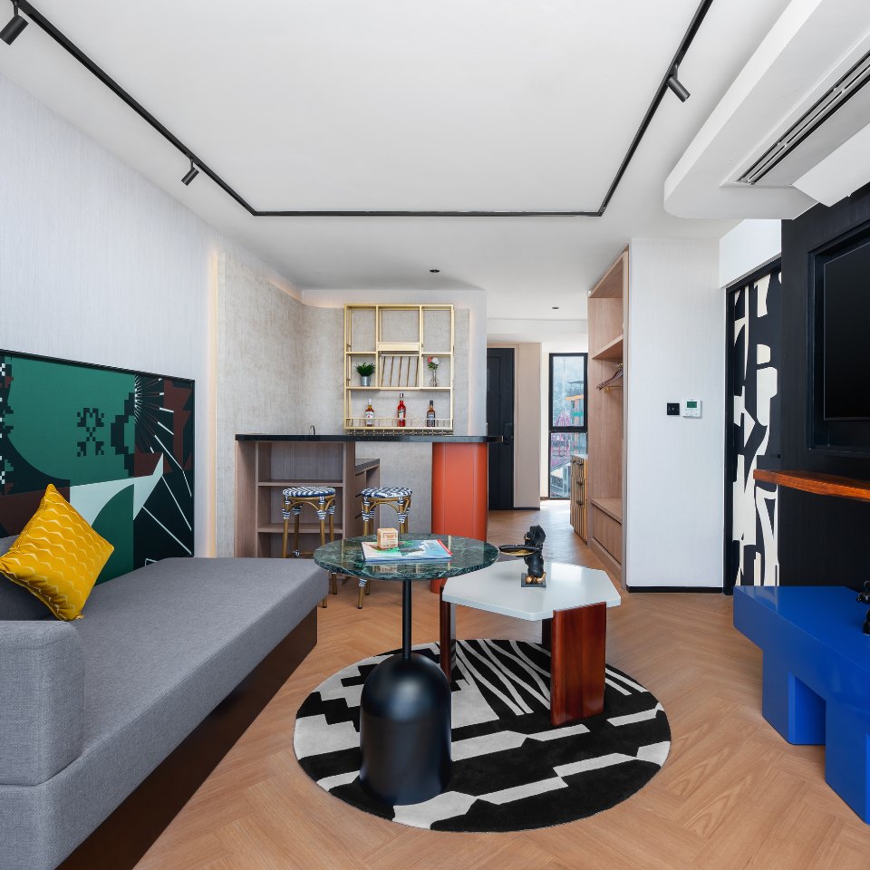 Modern living room with gray sofa, yellow pillow, patterned rug, two stylish coffee tables, bar stools, and open shelving with bottles.
