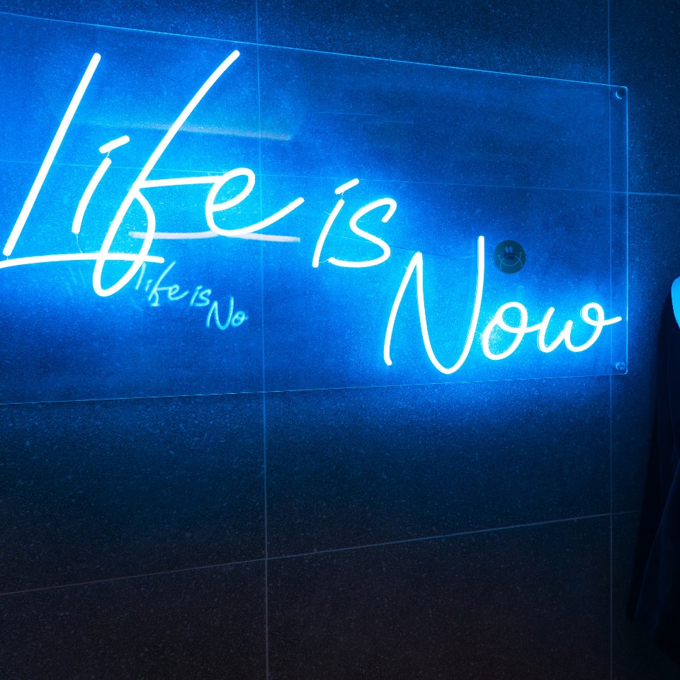 A glowing neon sign reads "Life is Now" in cursive, casting a vibrant blue light on a dark background, creating an inspiring and modern atmosphere.