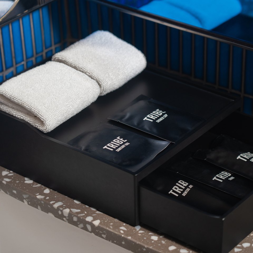 Black tray with neatly folded white towels and "TRIBE" branded sachets on a speckled countertop, creating a modern and organized look.