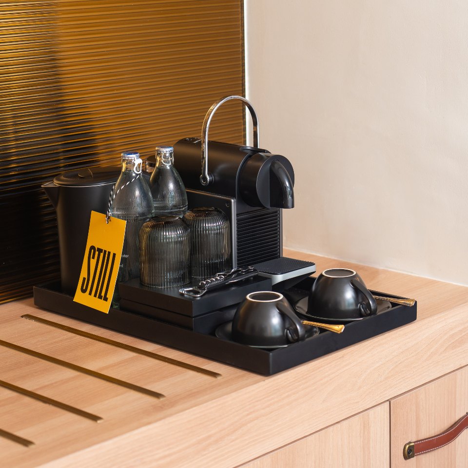 Sleek coffee station with a black espresso machine, glass bottles, cups, and a "STILL" tag on a wooden counter, creating a minimalist vibe.