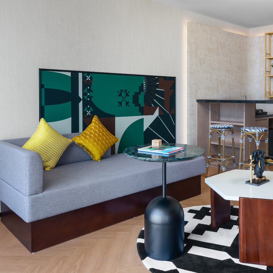 Modern living room with a gray sofa, yellow pillows, abstract wall art, marble side table, bar stools, and geometric rug.