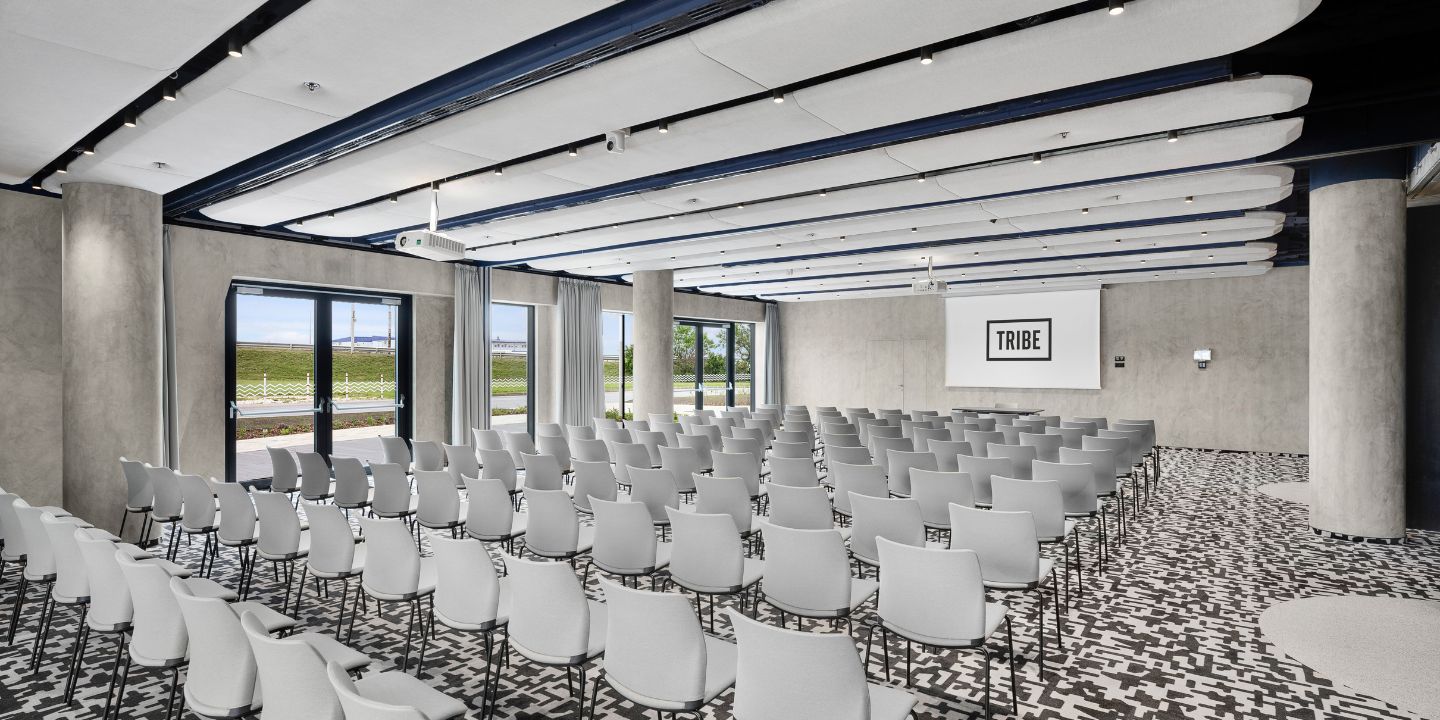 Spacious conference room with rows of white chairs, a geometric-patterned floor, large windows showing greenery, and a screen displaying "TRIBE."