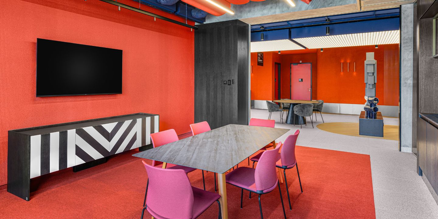 A modern meeting room with bold red walls and carpet, a large black TV, geometric sideboard, and a dark table surrounded by pink chairs. A second room with gray chairs and abstract sculptures is visible. The atmosphere is vibrant and contemporary.