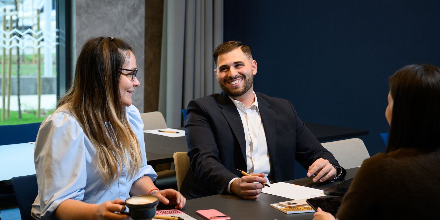 Three people in a modern office setting sit at a table engaged in a friendly discussion. One holds a coffee cup. All are smiling and appear engaged.