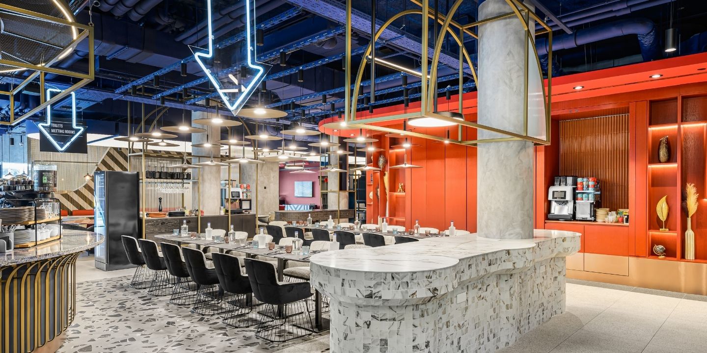 Modern restaurant interior with vibrant decor. Neon arrows hang from the ceiling, highlighting a long table with black chairs and a textured bar front. The red wall and varied lighting create an inviting, contemporary atmosphere.