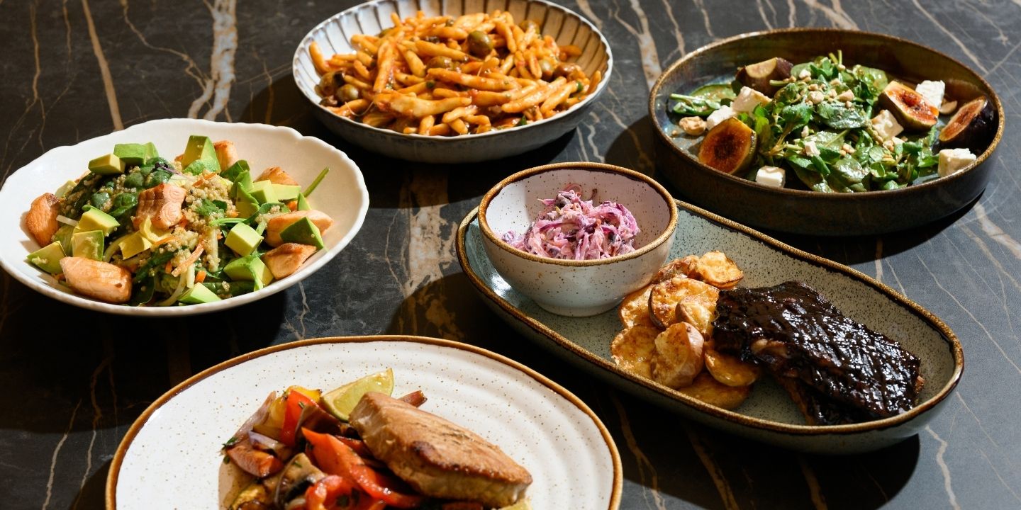 A variety of dishes on a dark marble table includes pasta, salads, ribs with plantains, fish, and roasted vegetables. The presentation is appetizing and vibrant.