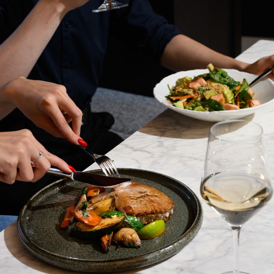 Two people dining at a marble table, one slicing meat with vegetables, another enjoying a salad. Wine glasses add a refined, relaxed ambiance.