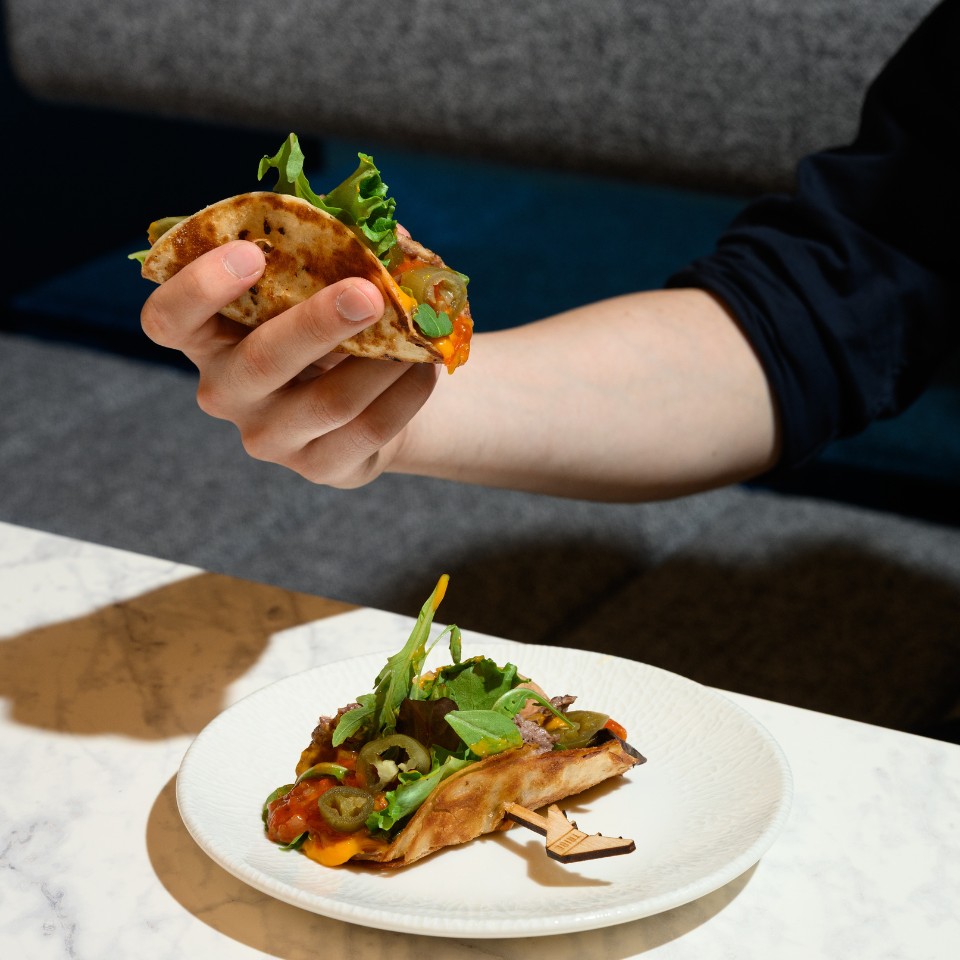 A hand holds a taco with lettuce, jalapeños, and melted cheese, above a marble table. Another similar taco is on a white plate, suggesting a casual dining setting.