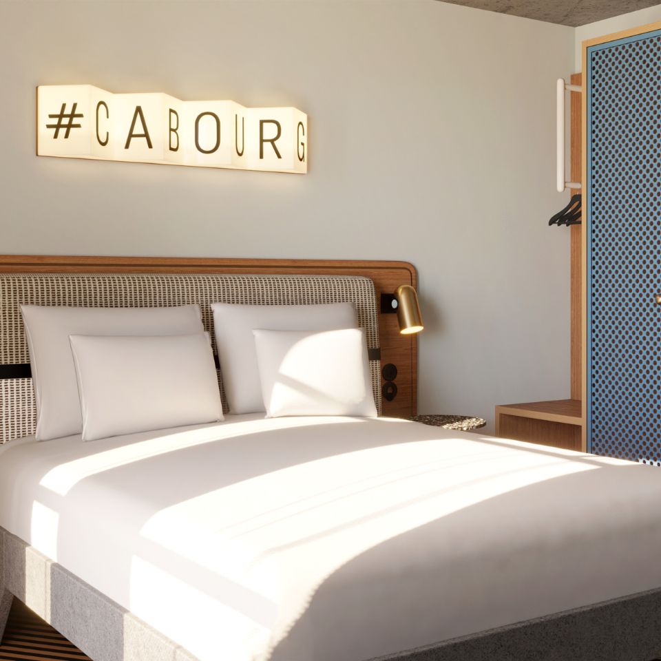 Sunlit modern hotel room with a double bed, white linens, a wooden headboard, wall lamp, and a blue patterned wardrobe.