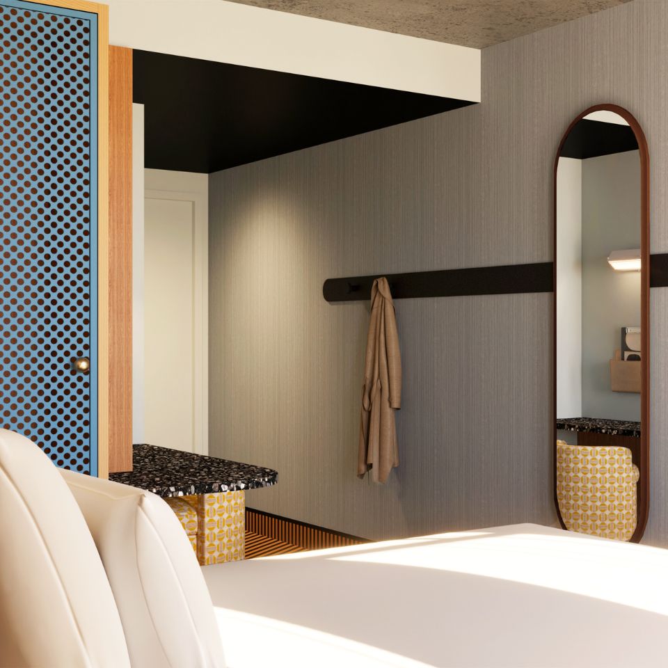 Modern hotel room corner with a beige coat hanging on a black wall hook, oval mirror, patterned chair, and blue polka-dot cabinet.