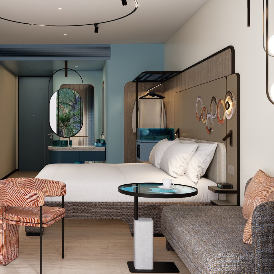 Cozy hotel studio suite corner featuring a grey fabric sofa, an orange geometric accent chair, a glass side table, and a plush king bed in the background.
