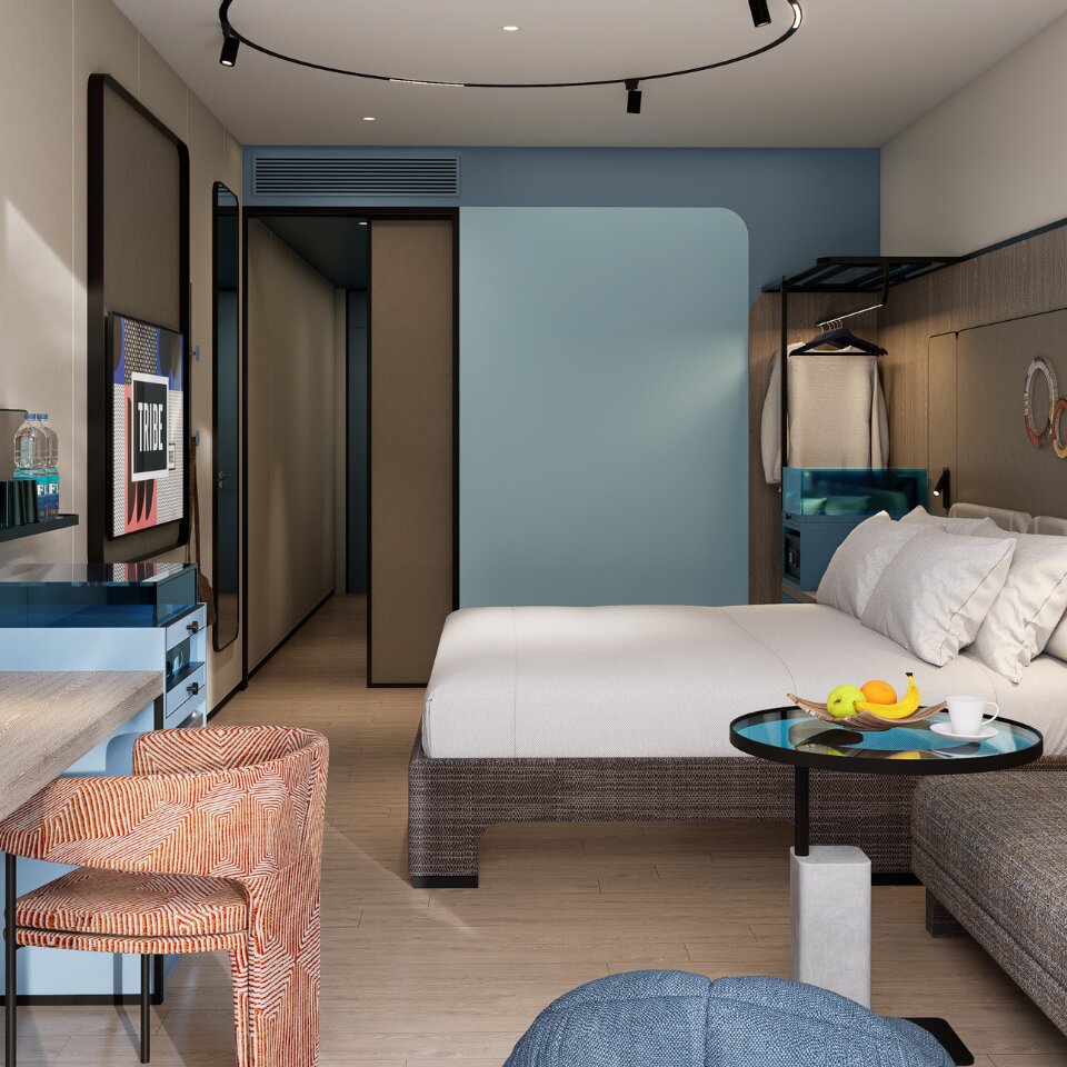 Cozy hotel studio suite corner featuring a grey fabric sofa, an orange geometric accent chair, a glass side table, and a plush king bed in the background.