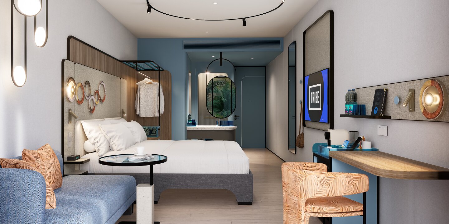 Stylish hotel studio suite showcasing a king bed, blue fabric lounge sofa, wooden desk with a textured armchair, and an open-concept washbasin vanity.