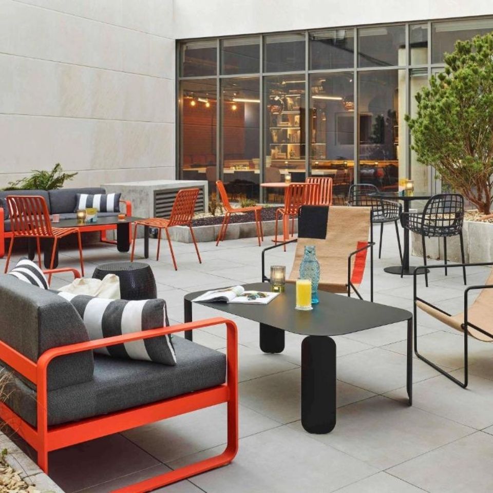 Spacious modern hotel terrace featuring contemporary grey sofas with bright orange metal frames, minimalist black tables, and large windows looking into the lobby.