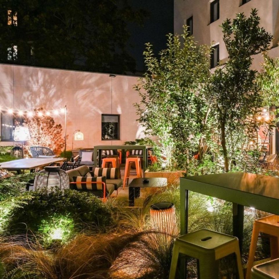 Cozy hotel courtyard patio at night featuring warm string lights, lush green plants, and modern seating with striped cushions and orange metal bar stools.