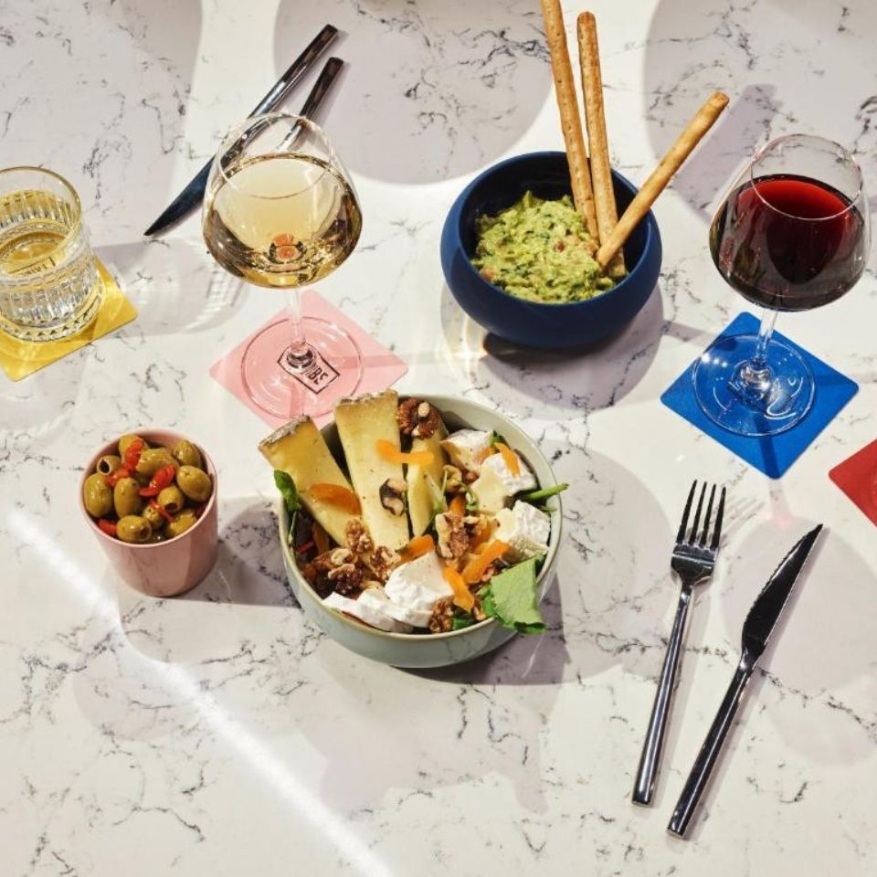 Flat lay of wine and appetizers on a white marble table, featuring a glass of white wine, red wine, a fresh cheese salad, green olives, and guacamole with breadsticks.