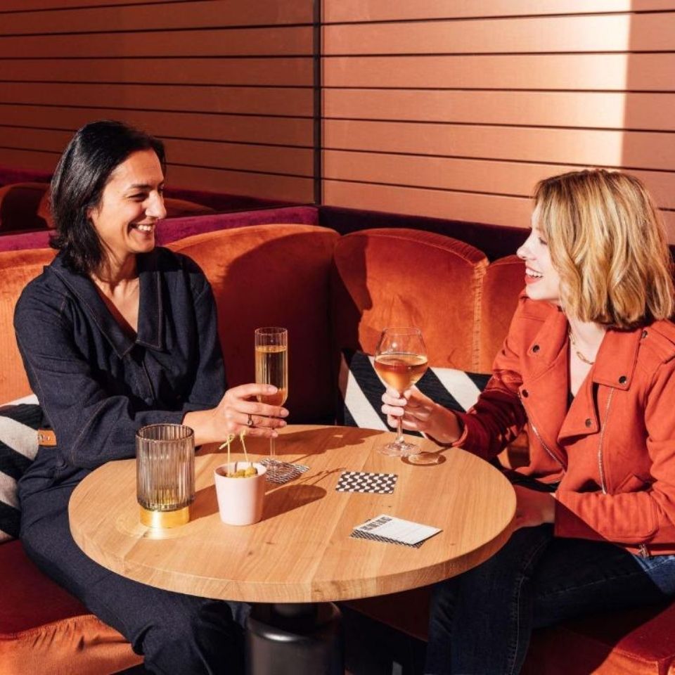 Two women smiling and chatting while enjoying glasses of champagne and wine in a cozy orange velvet booth at a modern hotel bar.