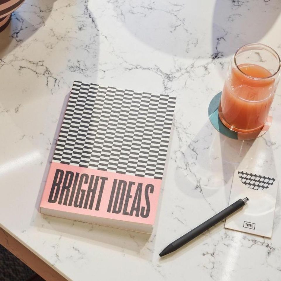 Flat lay of a 'Bright Ideas' checkered notebook, a black pen, and a glass of fresh juice on a marble desk at a modern hotel co-working space.
