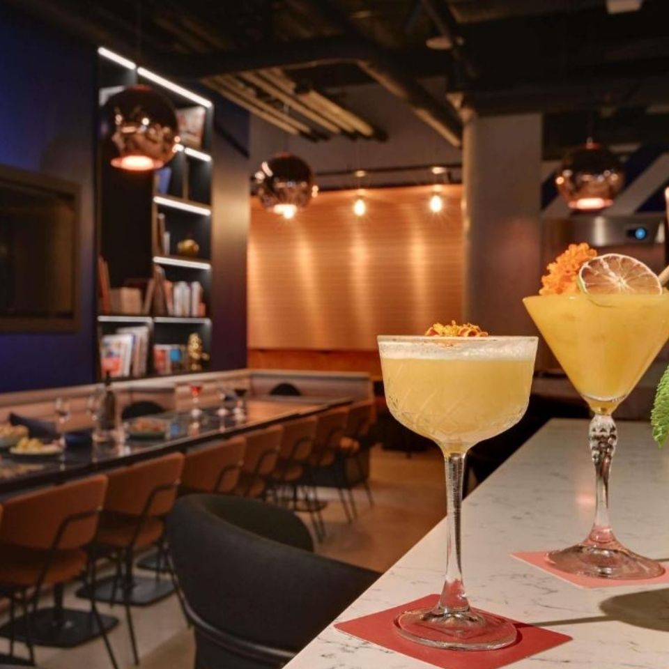 Two vibrant yellow craft cocktails garnished with dried citrus on a marble bar counter inside a dimly lit, modern hotel lounge with copper pendant lights.