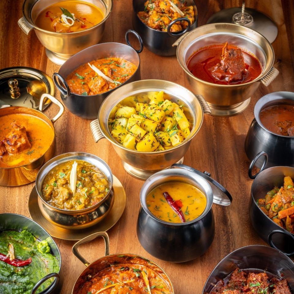 "A rich spread of traditional Indian curries, including Dal Tadka with a red chili, Jeera Aloo, and butter chicken, served in authentic brass and black metal bowls on a wooden table."