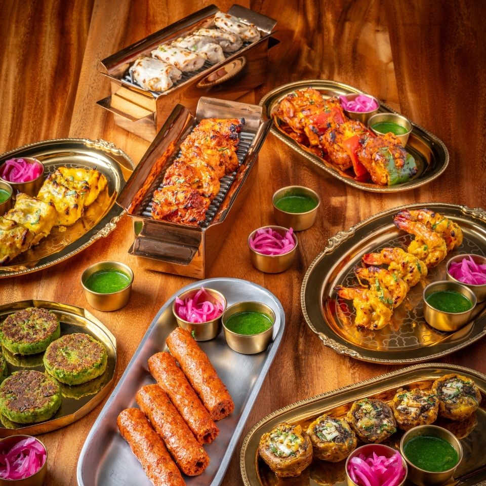"An assortment of Indian tandoori appetizers, featuring Seekh kebabs, grilled prawns, paneer tikka, and green hara bhara kababs, served on gold platters with mint chutney and pickled onions." 

