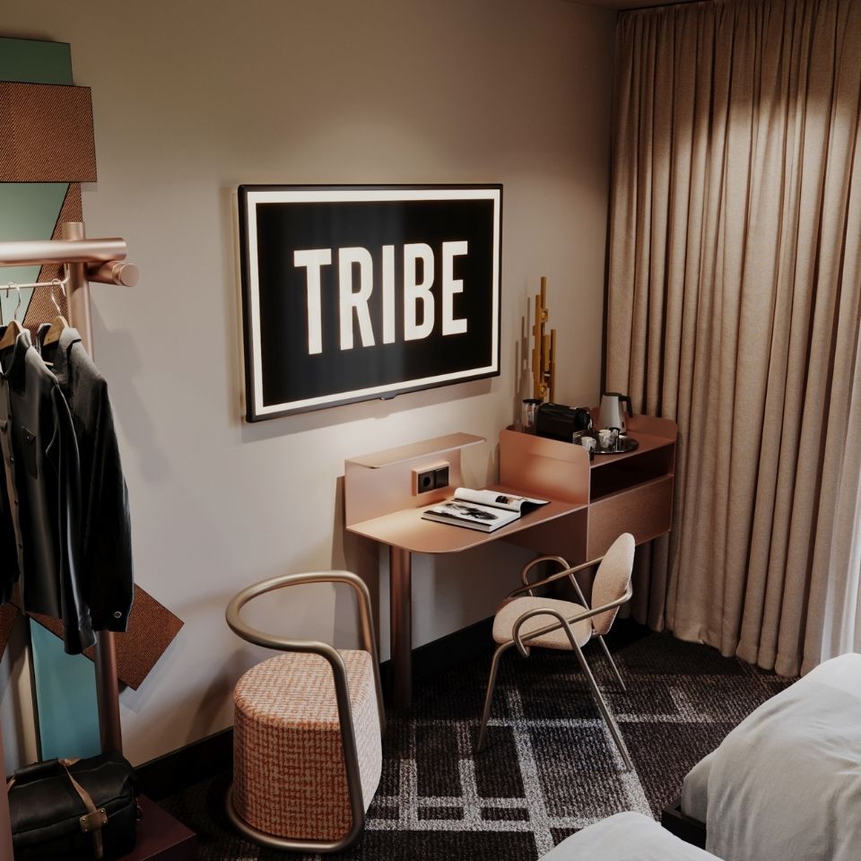 Sleek in-room workspace featuring a minimalist rose-gold desk, contemporary desk chairs, and a wall-mounted flat-screen TV displaying the 'TRIBE' logo.