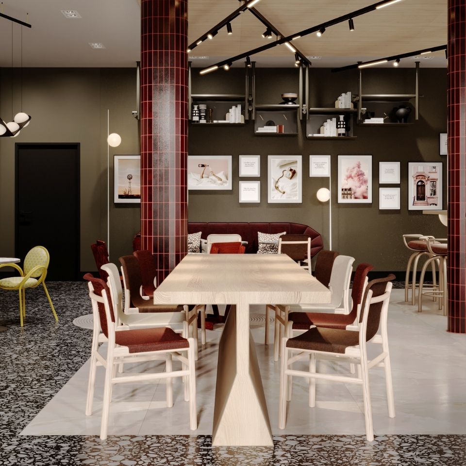 Large wooden communal co-working table in a modern hotel lobby, surrounded by contemporary chairs and red-tiled columns on a terrazzo tile floor.