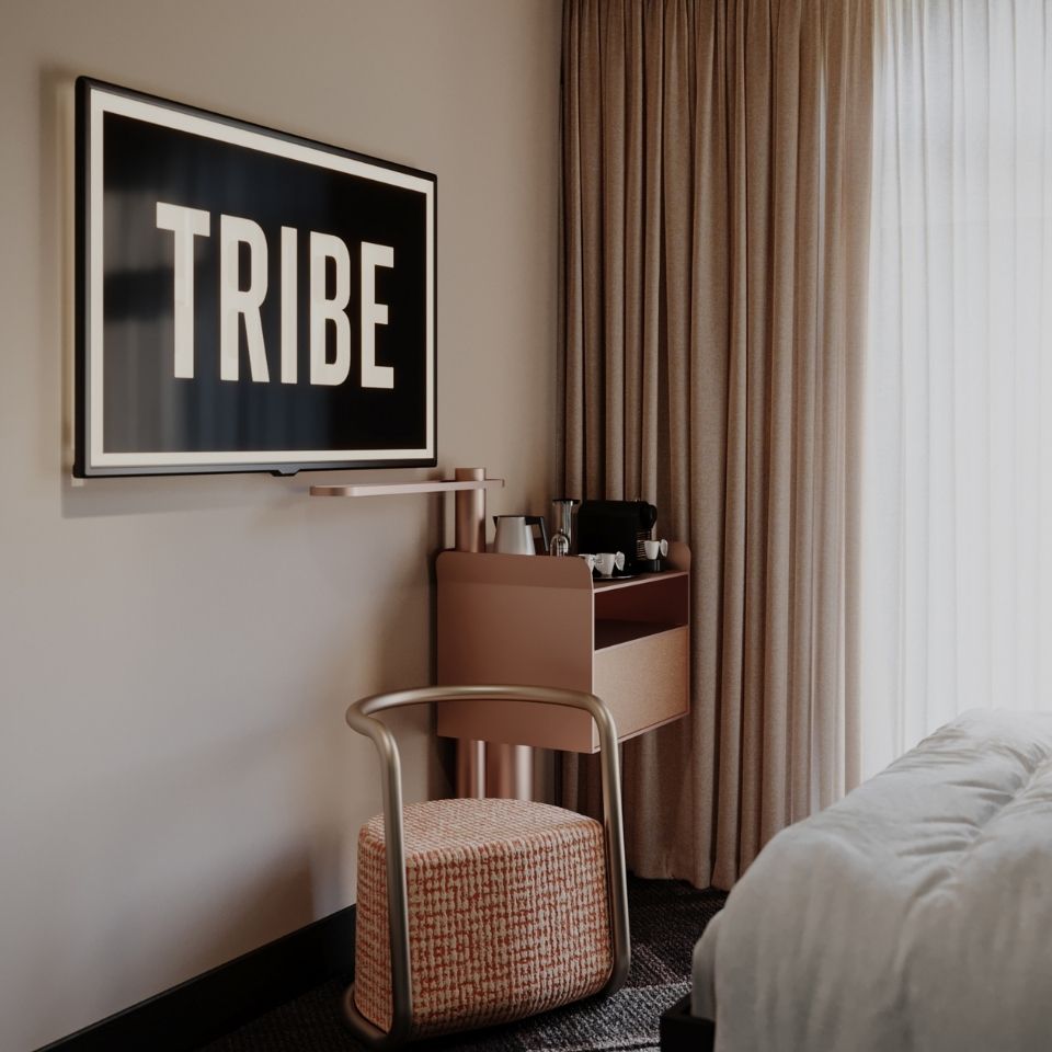 Side profile of a single hotel bed showcasing a multi-textured geometric headboard, a rose-gold cylindrical bedside table, and heavy cream-colored drapes.