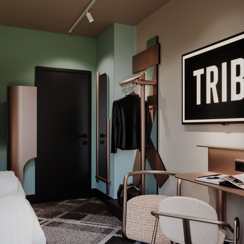 Hotel room entrance view showing a rose-gold open closet rack, a matching writing desk with a patterned cylindrical stool, and a flat-screen TV displaying TRIBE.