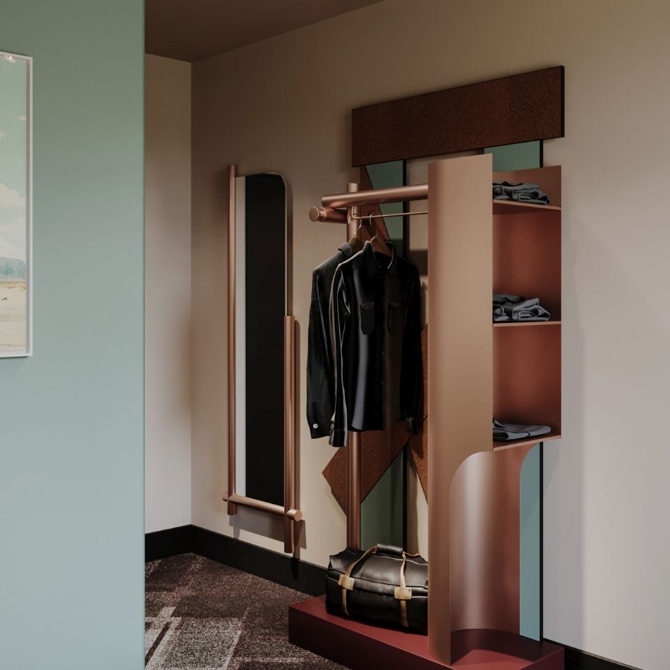 Minimalist open wardrobe closet in a hotel room, featuring a rose-gold metal clothing rack with hanging clothes, shelving, and a black duffel bag on a burgundy base.