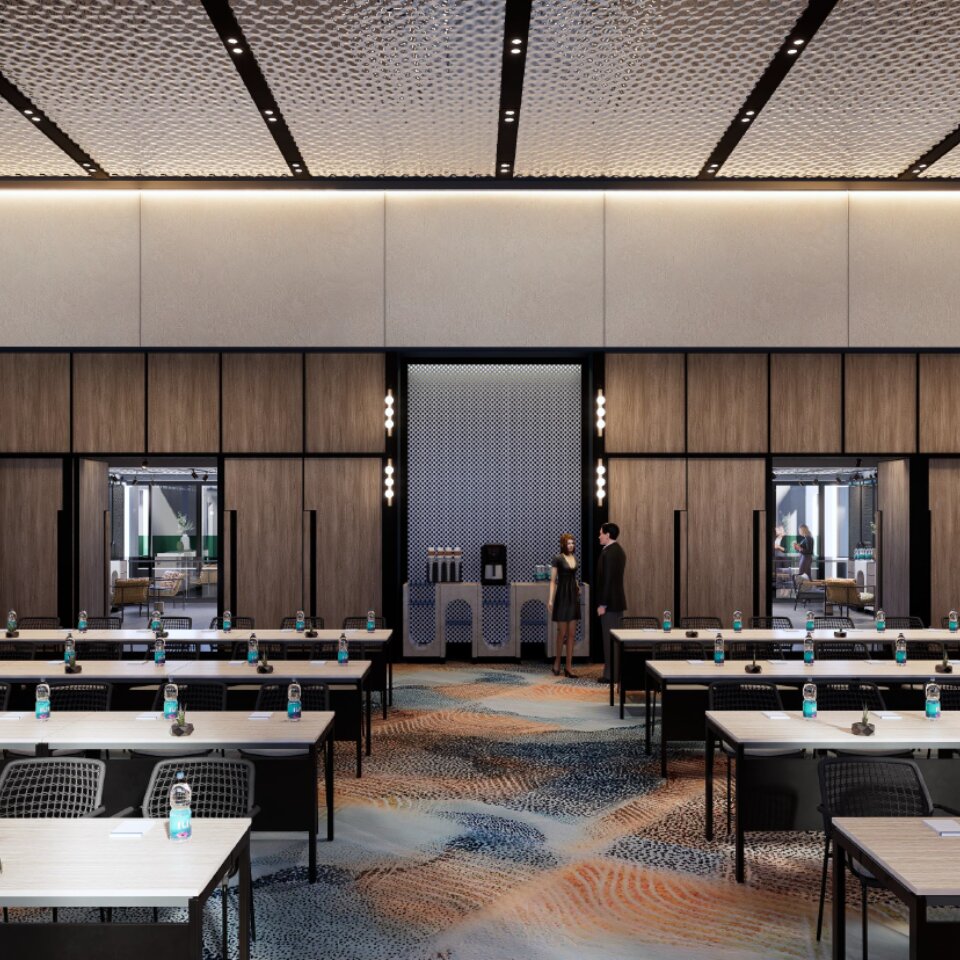 Large hotel conference room with classroom seating, long wooden tables with bottled water, vibrant patterned carpet, and wood-paneled walls.