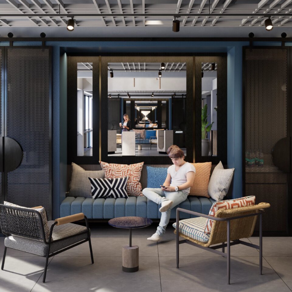 Cozy hotel lounge area with a blue upholstered built-in sofa, patterned accent pillows, modern armchairs, and a guest using a smartphone.