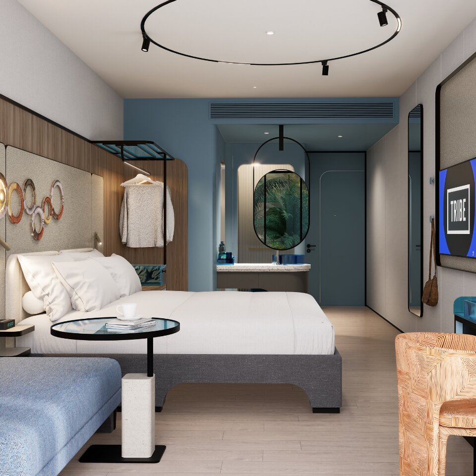 Sleek layout of a TRIBE hotel room featuring a king bed, blue lounge sofa, wooden desk, flat-screen TV, and contemporary lighting.