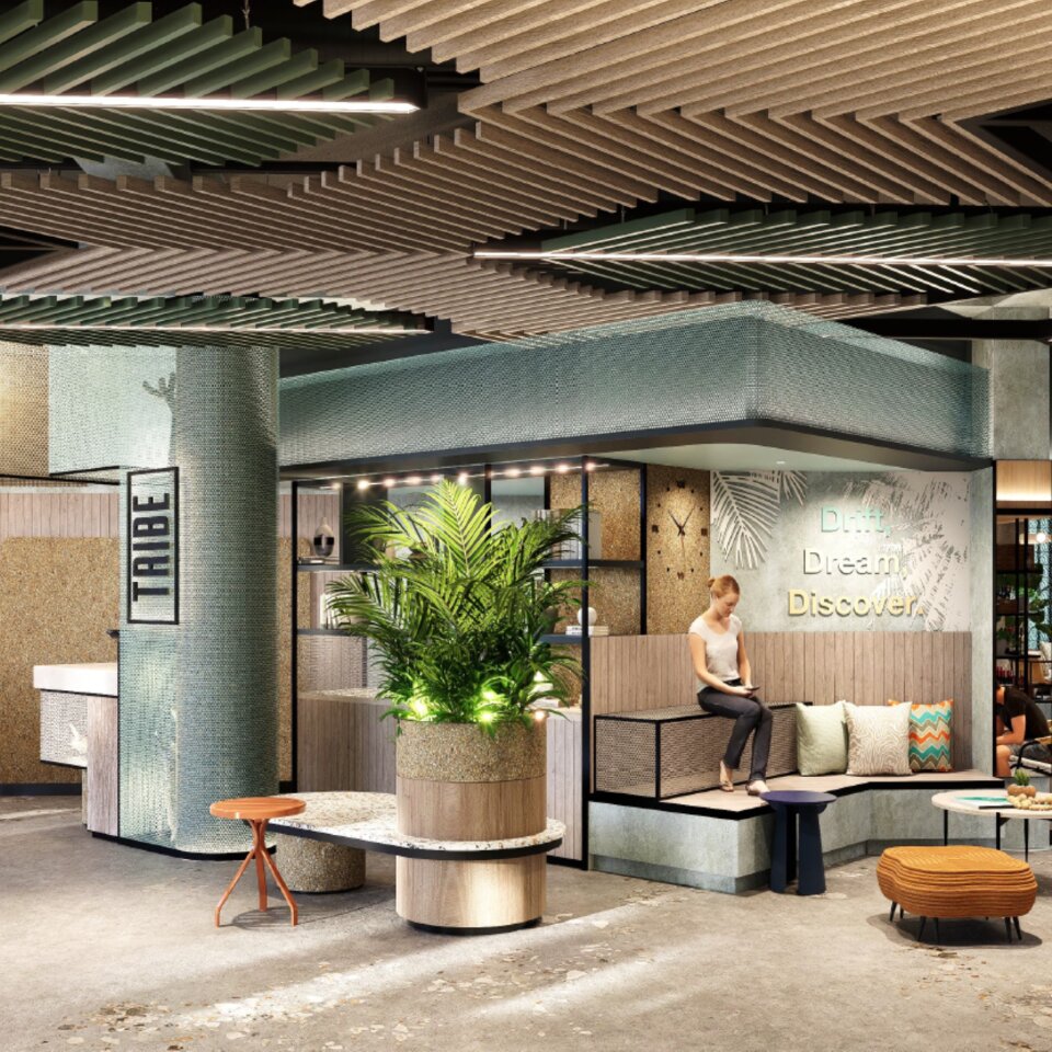 Modern hotel reception lobby with TRIBE branding, creative wooden slat ceiling, social seating corner, and lush potted green plants.
